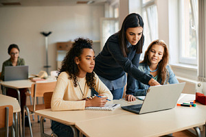 En lärare står vid två elever i ett klassrum och visar något på en datorskärm.