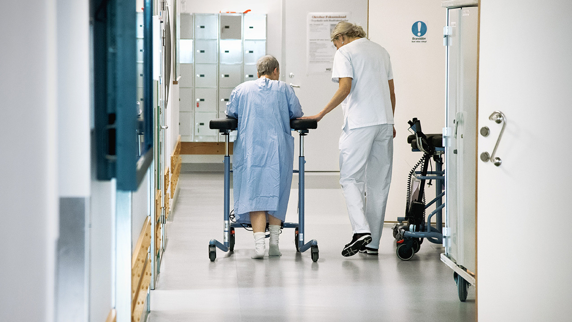 Vårdare hjälper gammal kvinna med rullator i vårdkorridor